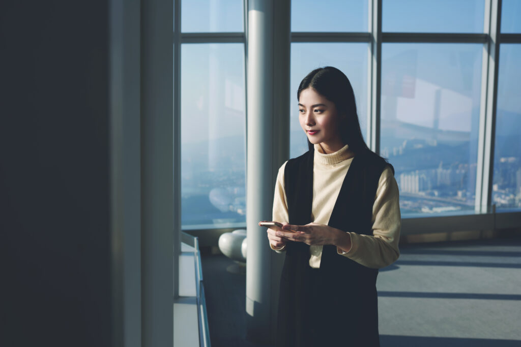 Asian hipster girl with trendy look is using mobile phone and looking on New York city from the window of last floor the skyscraper. Office worker of information technology company having work break
