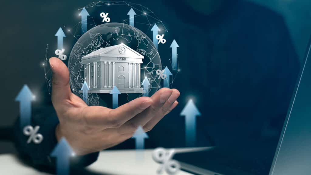 A hand holds a virtual bank surrounded by arrows and percentage signs, symbolizing rising interest rates and banking growth trends.