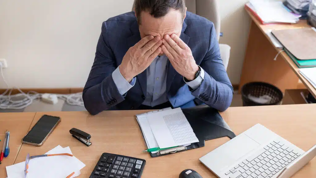 Small business owner experiences financial crisis, high costs, bank debt, money overspend concept. Man sitting at desk looks at laptop screen holding receipt feels desperate after calculating expenses
