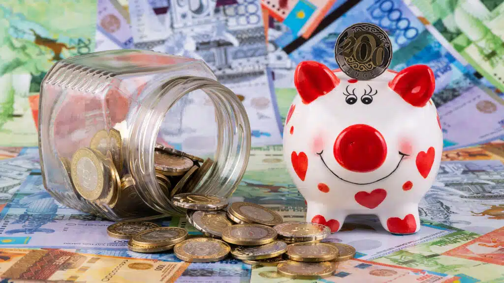 Glass jar, Kazakhstani coins of denomination of 100 and 200 tenge and a piggy bank against the background of paper Kazakhstani tenge of different denominations