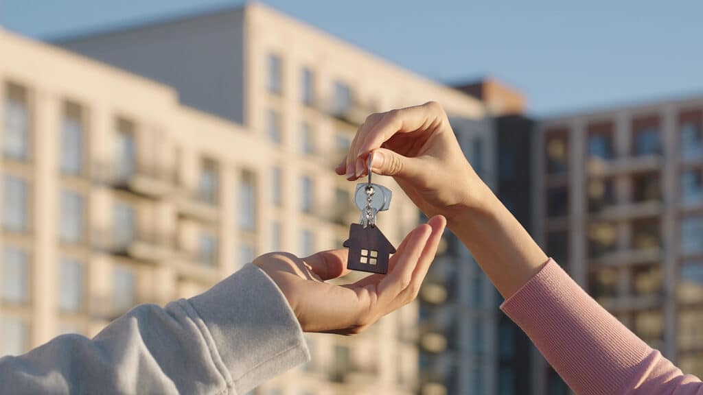 Close up of female real estate owner handing over house key with home shaped keychain to young man outdoors on background of modern apartment building. New home, first time buyer, and property deal.