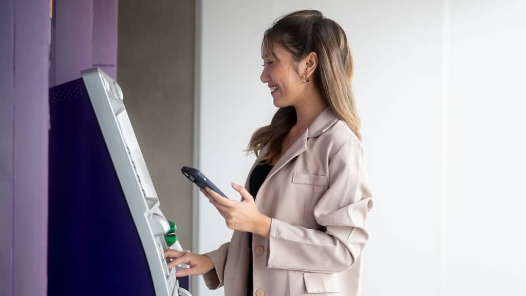 Smiling asian woman standing holding phone in front of an atm machine waiting for a withdraw money or cash. Transaction, Travel card, Bank Advertising.